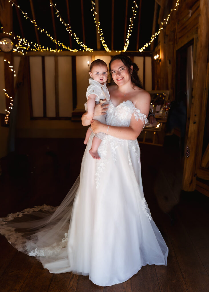 A smiling bride in a white wedding dress holds a baby in a rustic venue decorated with string lights.