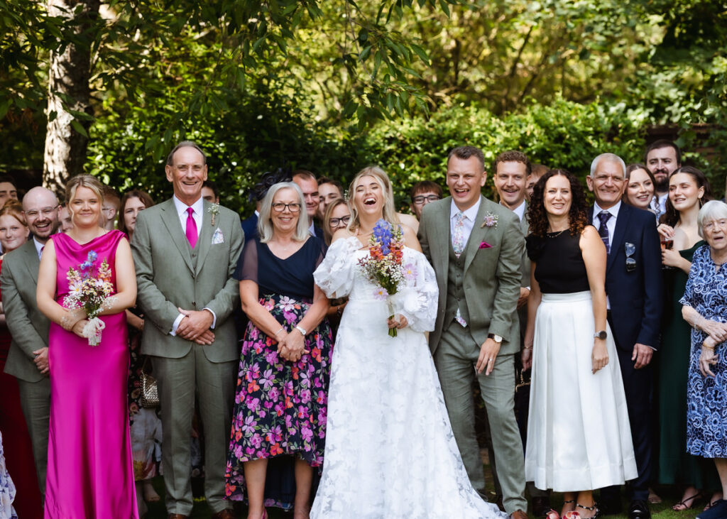 A large group of people, including a bride in a white dress and others in formal attire, stand outdoors smiling and laughing, surrounded by greenery.