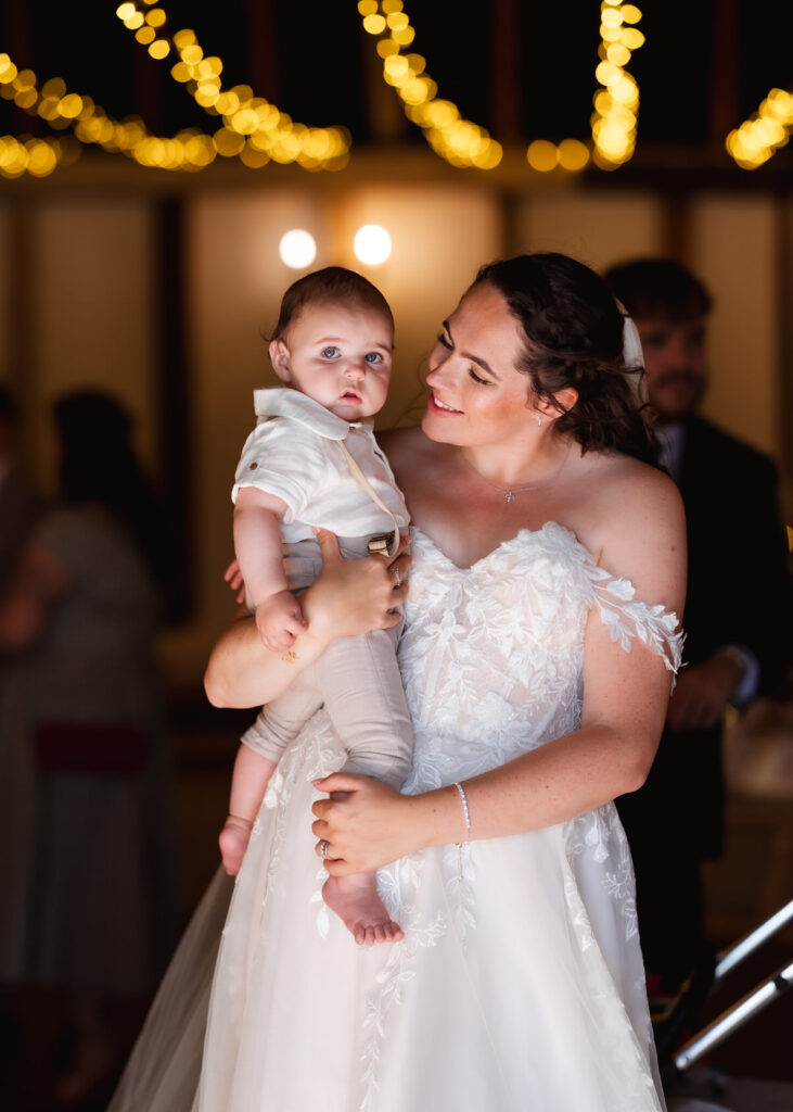 A woman in a white wedding dress holds a baby in her arms, both looking content. Warm yellow string lights hang in the background, creating a festive atmosphere.