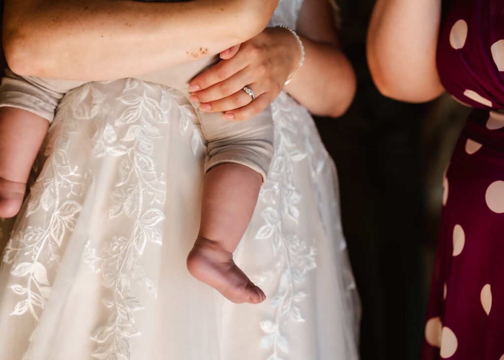 A close-up of a bride in a white lace gown holding a baby, with another person in a maroon polka dot dress beside her. The brides engagement ring is visible on her hand.