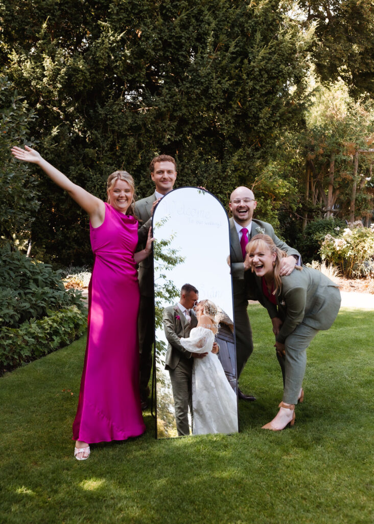 Four people pose joyfully around a tall mirror outdoors; the mirror reflects a bride and groom kissing, surrounded by lush greenery and sunlight.