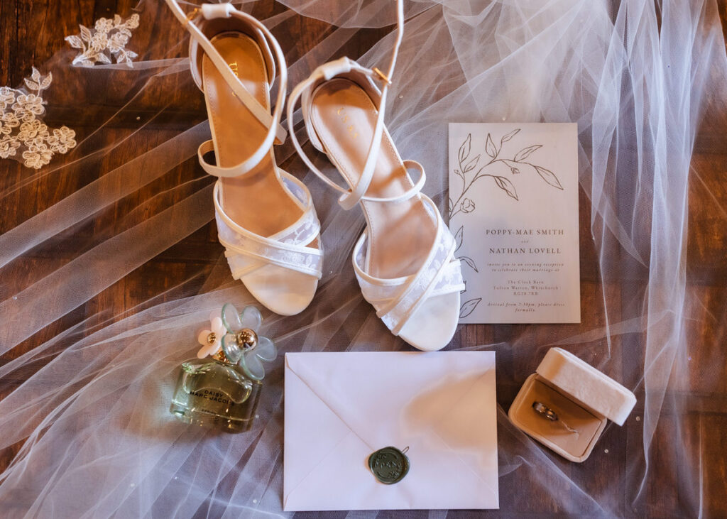 White heels, perfume, a lace hairpiece, a wedding invitation, an envelope with a wax seal, and a ring in a box are arranged on sheer fabric, suggesting a bridal preparation scene.