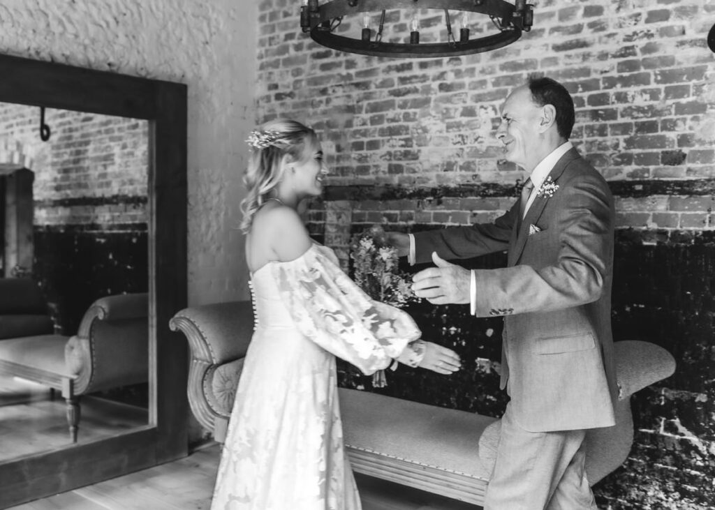 A woman in a wedding dress and a man in a suit smile and move toward each other with open arms, preparing to hug in a rustic room with exposed brick walls.