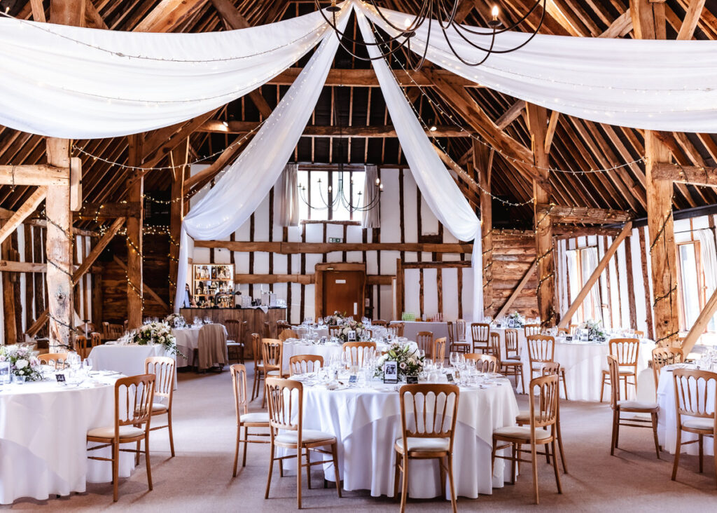 A rustic barn decorated for a wedding, featuring round tables with white tablecloths, elegant place settings, flower centerpieces, wooden chairs, draped white fabric, and string lights hanging from the wooden beams.