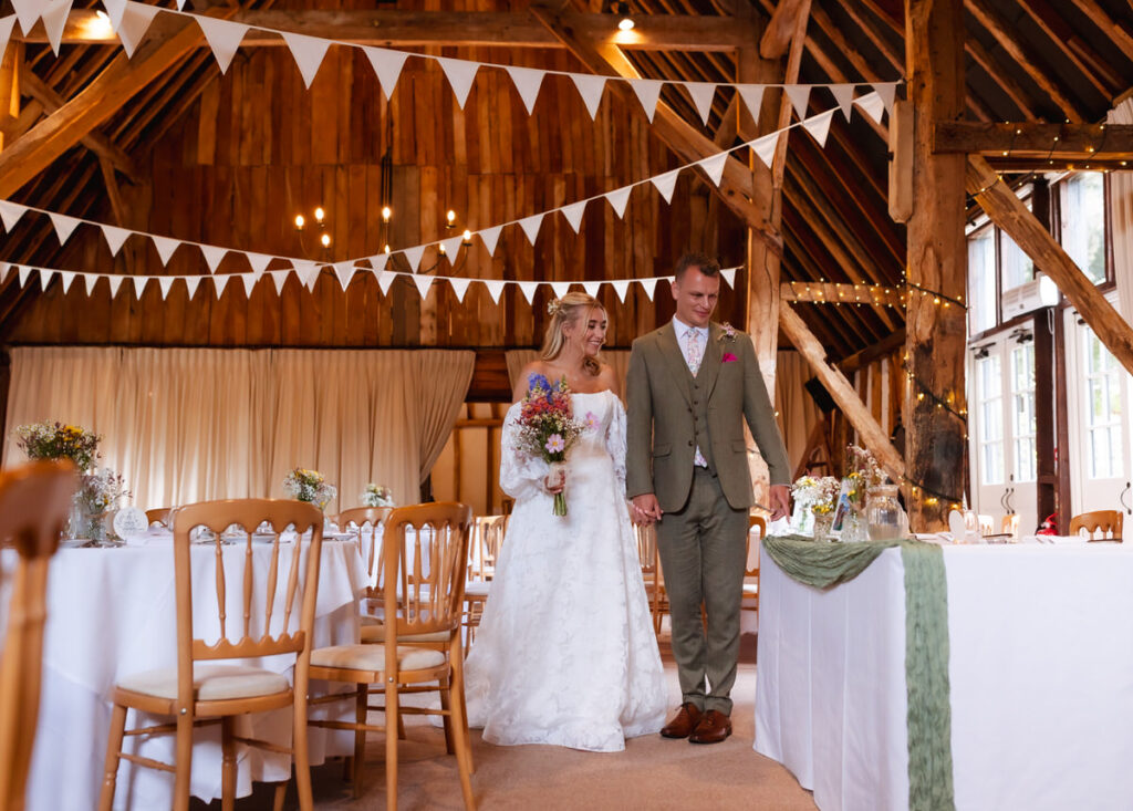 A bride and groom, holding hands, walk through a rustic barn decorated with white bunting, string lights, and wooden chairs, with tables set for a wedding reception.