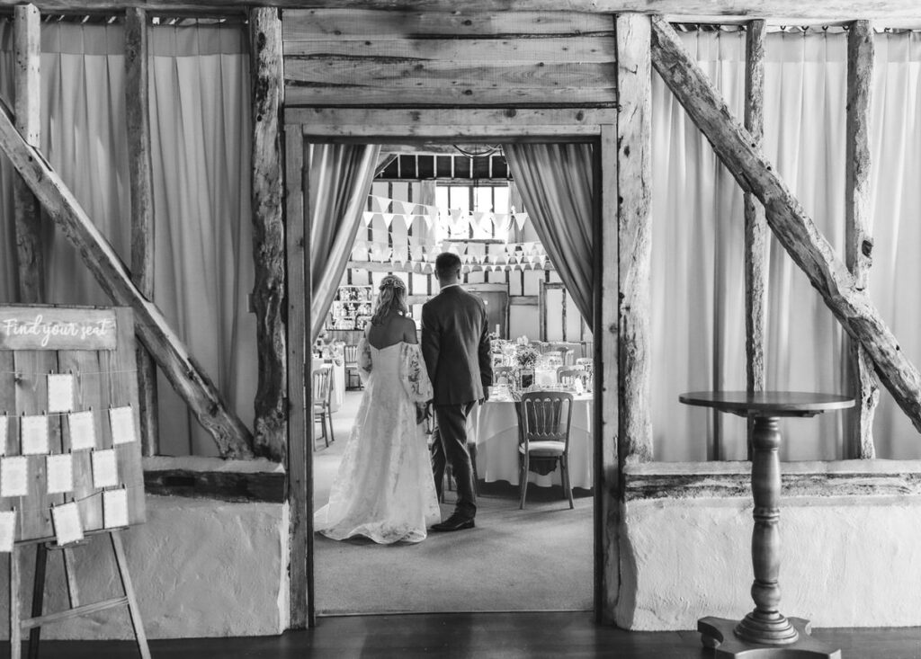 A bride and groom, seen from behind, enter a rustic wedding reception hall decorated with tables and string lights. The scene is framed by wooden beams and curtains.
