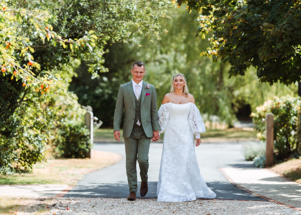A groom in a green suit and a bride in a white dress walk hand in hand down a tree-lined path, smiling outdoors on a sunny day.
