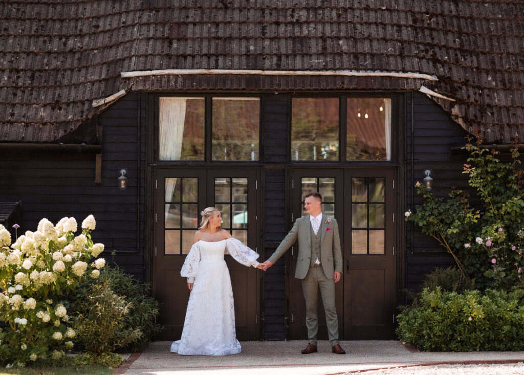 A bride and groom hold hands and face each other outside a rustic dark wooden building, surrounded by green bushes and white flowers.