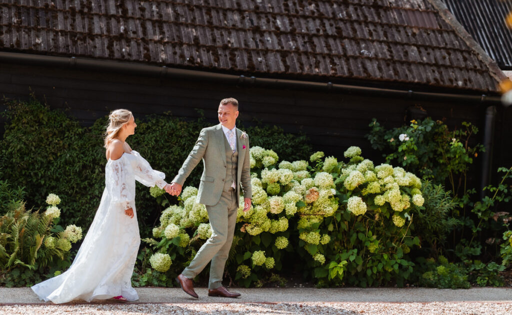 A bride and groom walk hand in hand outside, smiling at each other, with green hydrangea bushes and a rustic dark building in the background.