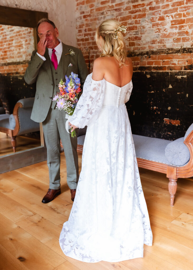 A man in a suit wipes away tears while seeing a woman in a white wedding dress holding a colorful bouquet. They stand on a wooden floor with brick walls and vintage benches in the background.