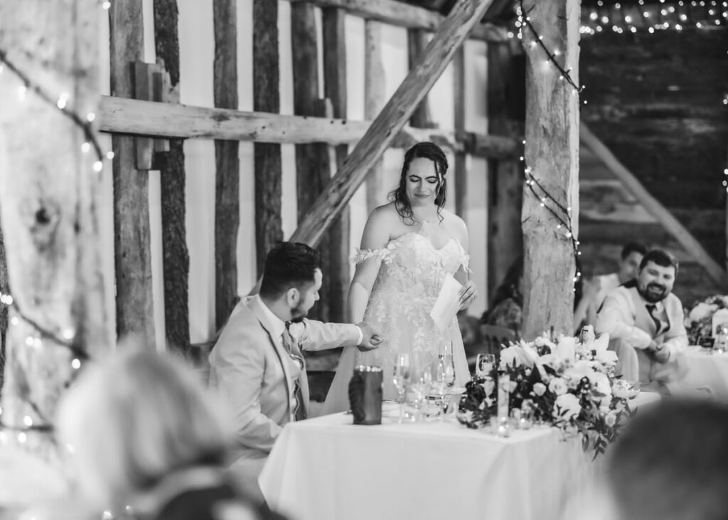 A bride in a white dress stands smiling with a piece of paper, while a seated man in a suit gently holds her hand. The scene is decorated with flowers and string lights in a rustic setting.