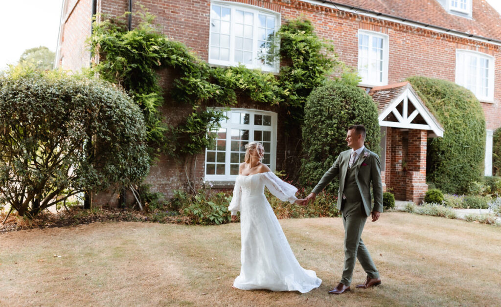 A bride and groom hold hands and smile while walking on a lawn in front of a brick house with large windows and greenery.