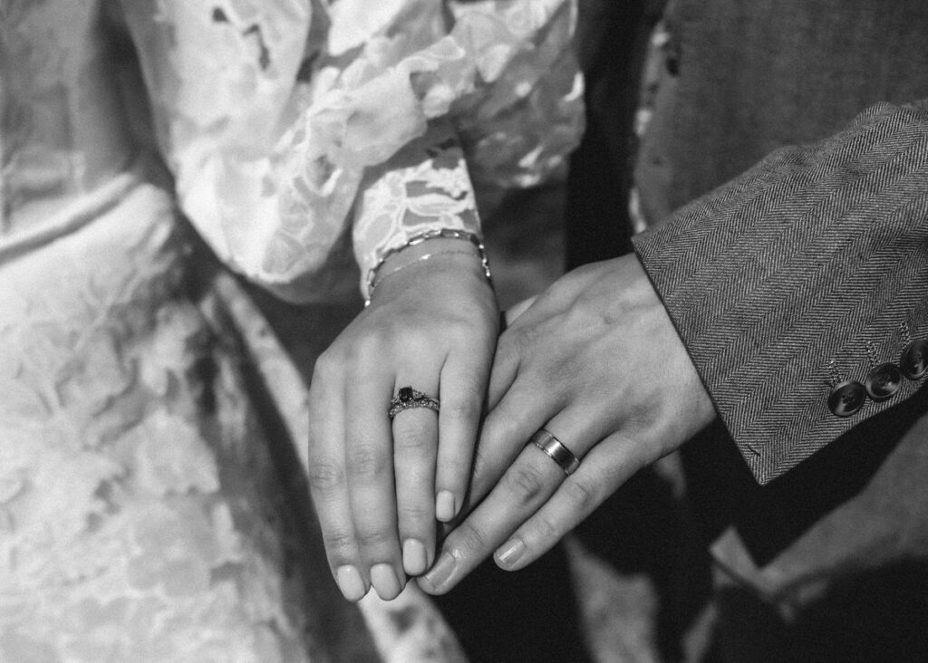 Two hands with wedding rings rest side by side. One hand has a delicate lace sleeve and an engagement ring; the other has a plain wedding band and a suit sleeve.