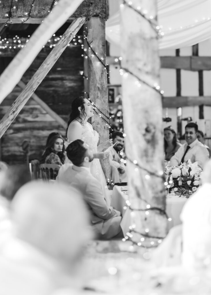 A bride stands and speaks at a wedding reception in a rustic, decorated venue, surrounded by guests and twinkling string lights.