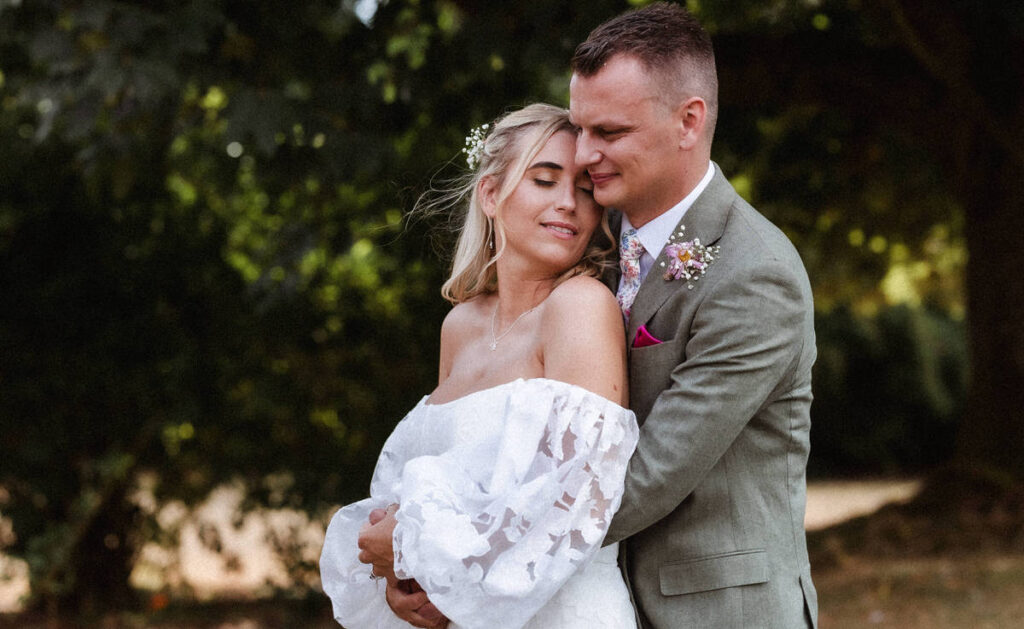 A bride in a white off-shoulder dress and a groom in a light suit embrace outdoors, both with closed eyes, standing under trees with soft natural light.