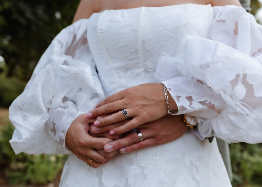 A close-up of a couple embracing, with both wearing rings and the person in front dressed in a white, off-the-shoulder, floral wedding gown. The hands are gently intertwined at the waist.