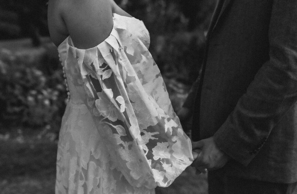 A black-and-white photo showing two people holding hands. One wears a floral, off-the-shoulder dress, while the other is in a suit. Only their torsos and hands are visible.