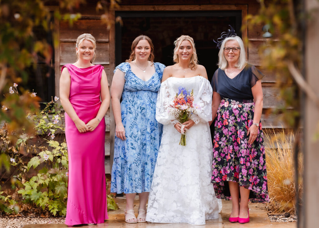 Four women stand side by side outdoors, smiling at the camera. One wears a white wedding dress, holding a bouquet, while the others wear colorful dresses. The background features greenery and a wooden building.