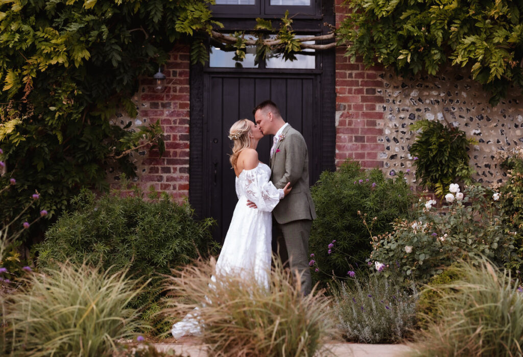 A bride and groom stand close, touching noses and holding hands, in front of a rustic brick building with lush greenery and flowers around them.