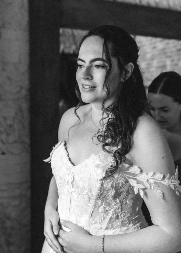 A bride in an off-the-shoulder lace wedding dress stands indoors, smiling softly. Another woman is visible behind her, slightly out of focus. The photo is in black and white.