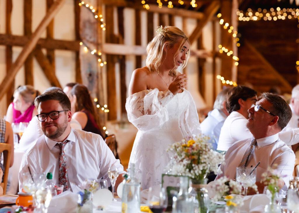 A woman in a white off-shoulder dress stands smiling and talking to two seated men at a festive event, with rustic wooden walls and string lights in the background.