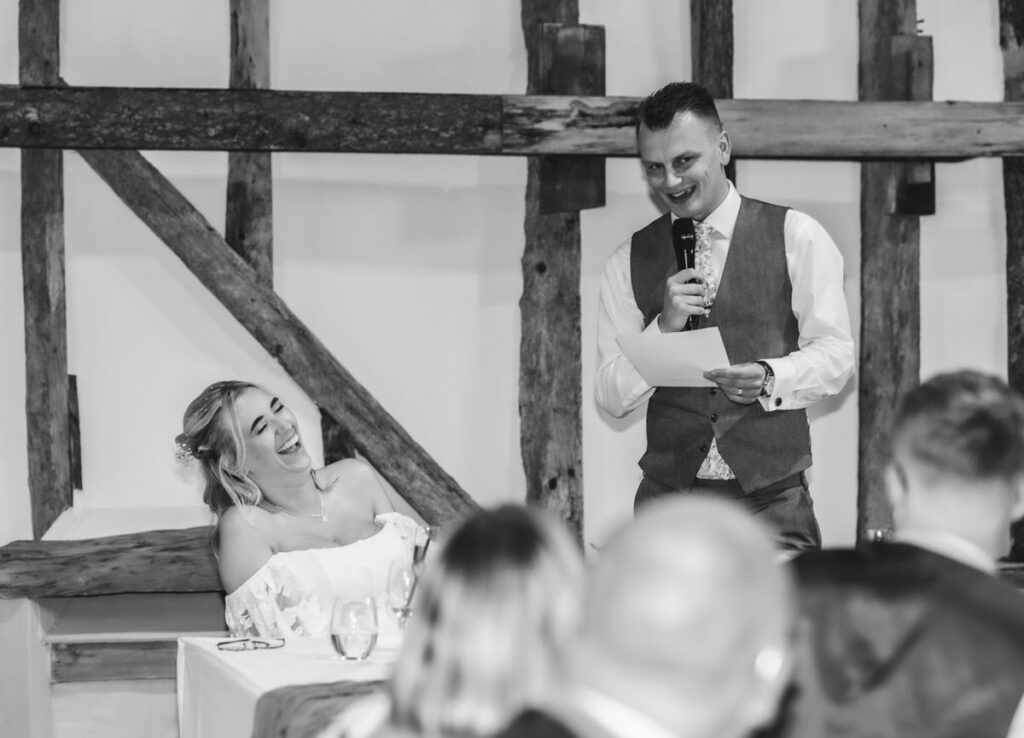 A man in a vest and tie gives a speech with a microphone while a woman in a white dress laughs at a table during a wedding reception. Other guests are visible in the foreground.