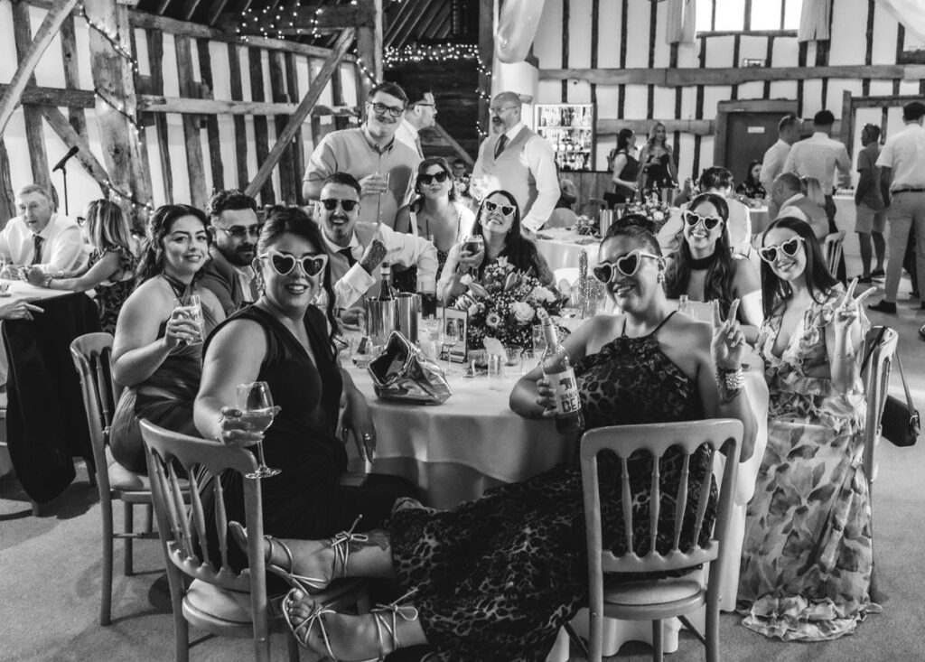 A group of people in fun sunglasses sit around a decorated table at a lively indoor event, smiling, posing, and making peace signs. The barn-like venue is filled with guests and string lights.