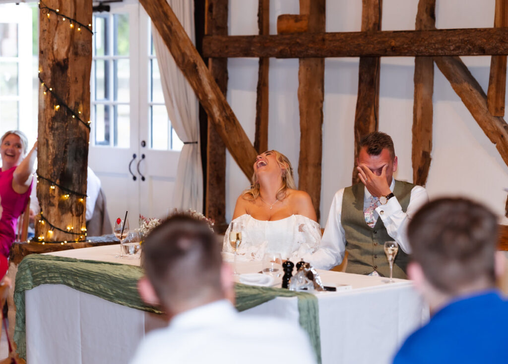 A bride and groom sit at a table laughing heartily during a wedding speech. Wooden beams and fairy lights decorate the venue. Guests in the foreground enjoy the moment.