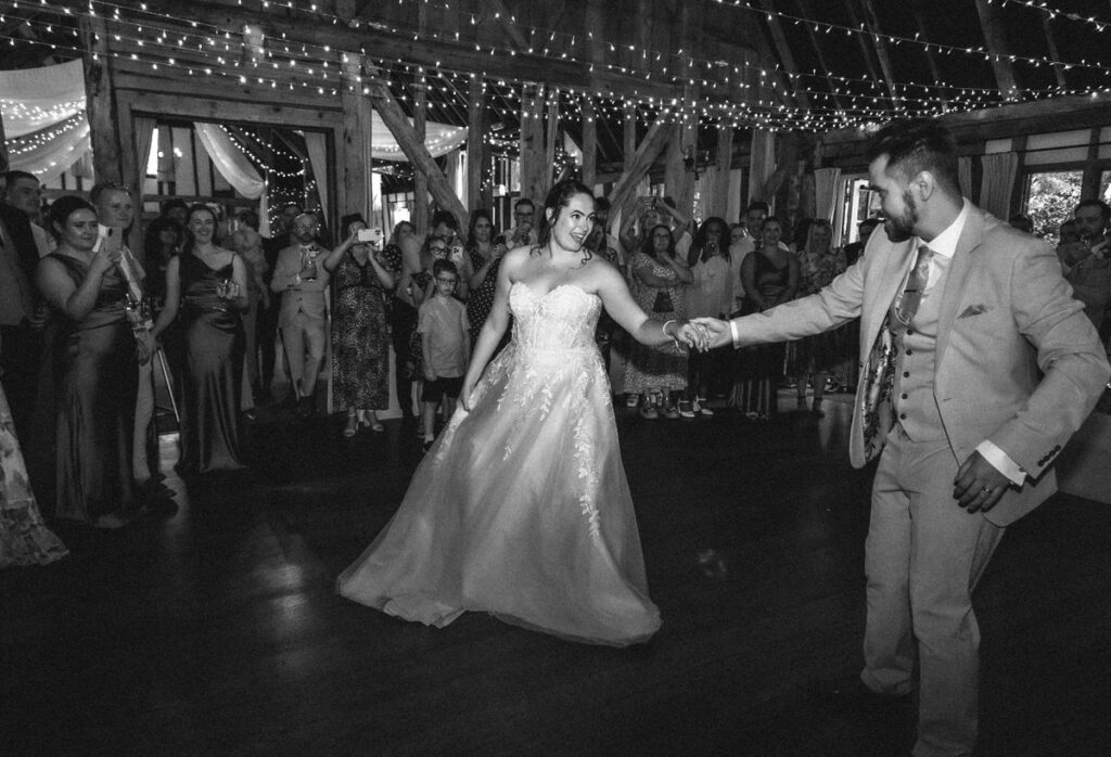 A bride and groom share their first dance in a rustic venue with string lights, surrounded by guests watching and celebrating.