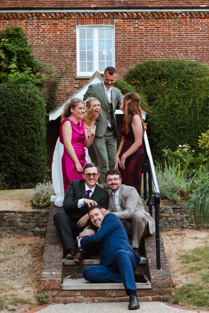 Seven adults in formal wear pose playfully on outdoor steps in front of a brick house, smiling and laughing. Three sit on the lower steps while four stand above, some holding drinks. Green bushes and a garden surround them.