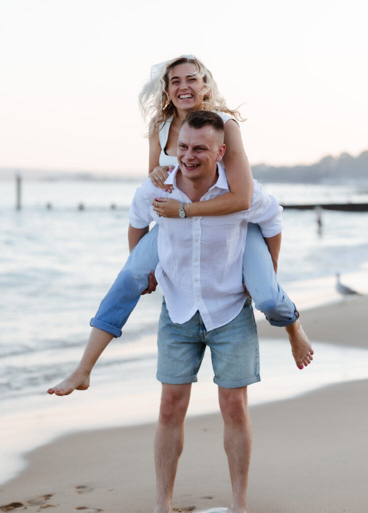 A smiling woman in light clothes is on a mans back, both laughing on a sandy beach near the water. The man wears a white shirt and shorts. The scene is bright and relaxed, with gentle waves in the background.
