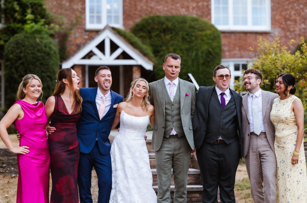 A group of seven people, including a bride in a white wedding dress and a groom in a grey suit, pose playfully together outdoors in front of a brick building.
