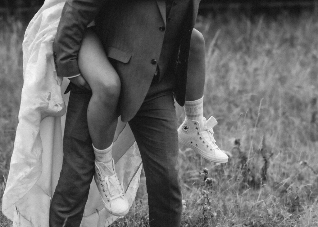 A groom in a suit carries a bride piggyback style through a grassy field. The bride wears a dress, white socks, and sneakers labeled BRIDE. The image is in black and white.