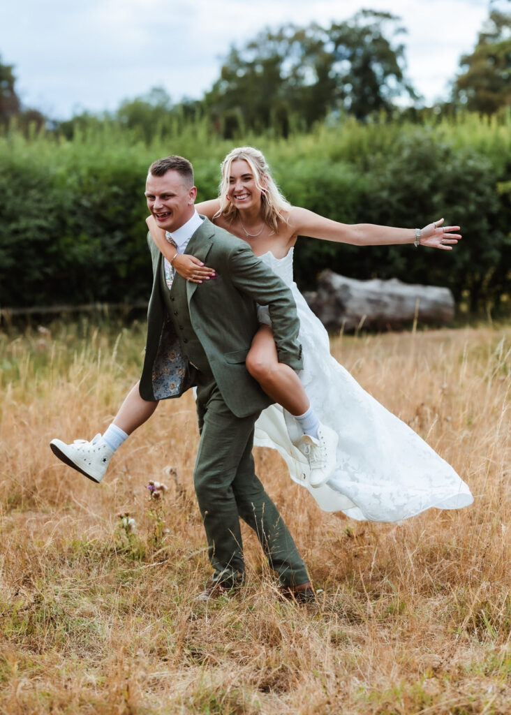 A groom in a green suit gives a piggyback ride to a smiling bride in a white dress and sneakers. They are outdoors in a grassy field with trees in the background.