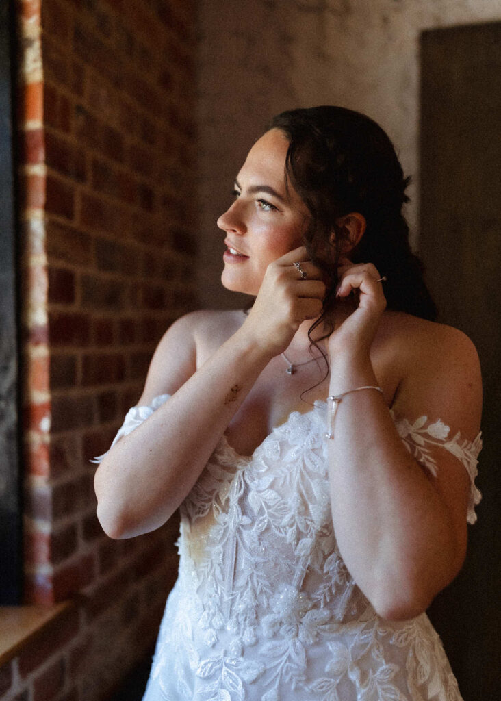 A bride in an off-shoulder white lace gown stands by a brick wall, smiling softly as she adjusts an earring, with warm natural light illuminating her face.