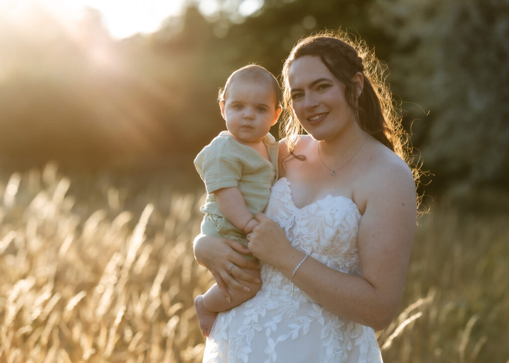 A woman in a white dress holds a baby in a field of tall grass, both looking at the camera with sunlight shining behind them.