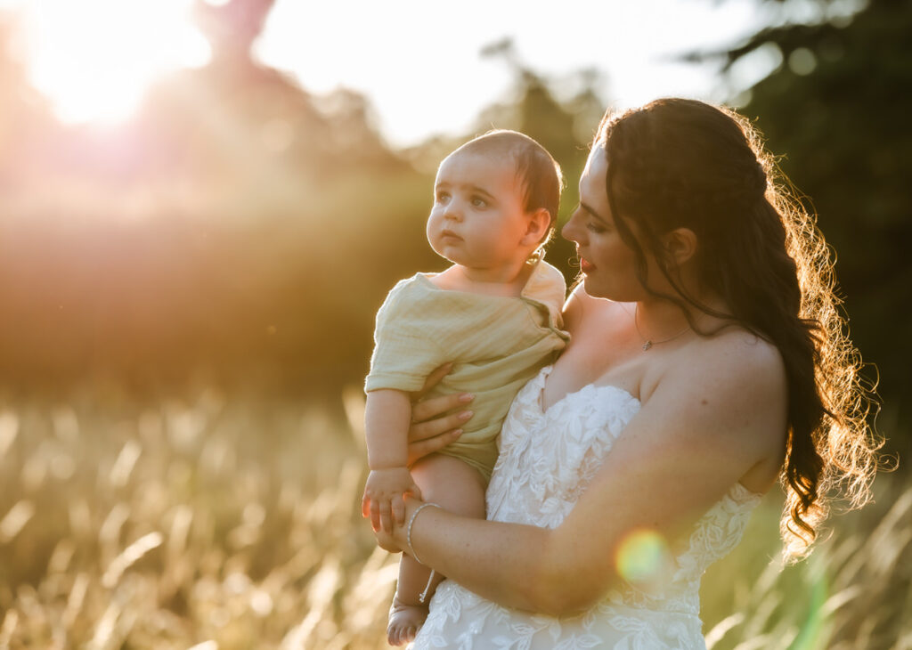 A woman in a white dress holds a baby in a field during golden hour, with sunlight shining through grass and trees in the background.
