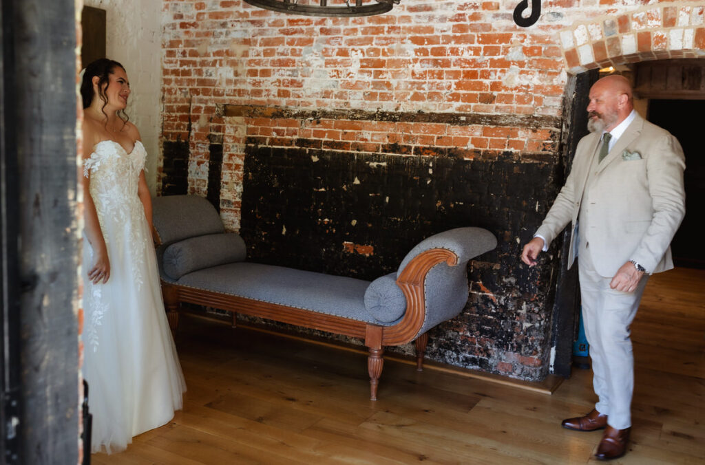 A bride in a white dress smiles at a man in a light suit, both inside a rustic room with exposed brick walls and a wooden floor. The man looks surprised and happy.