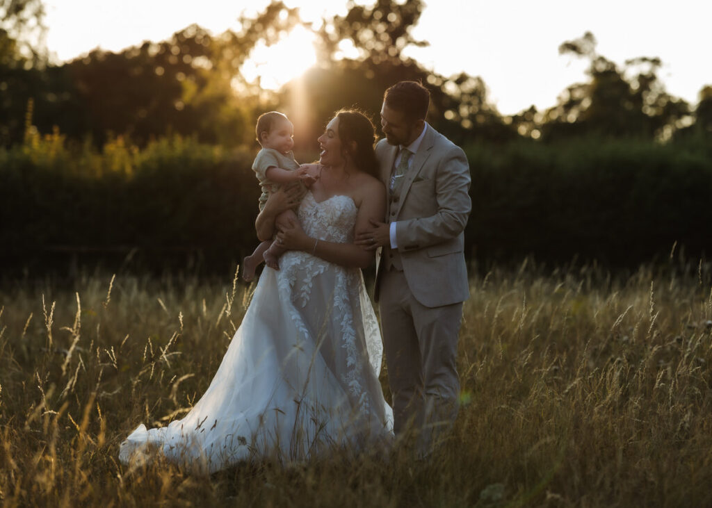 A bride and groom stand in a sunlit field, smiling and holding a baby together. The bride wears a white gown, the groom a tan suit, and the scene is surrounded by tall grass and trees in the background.