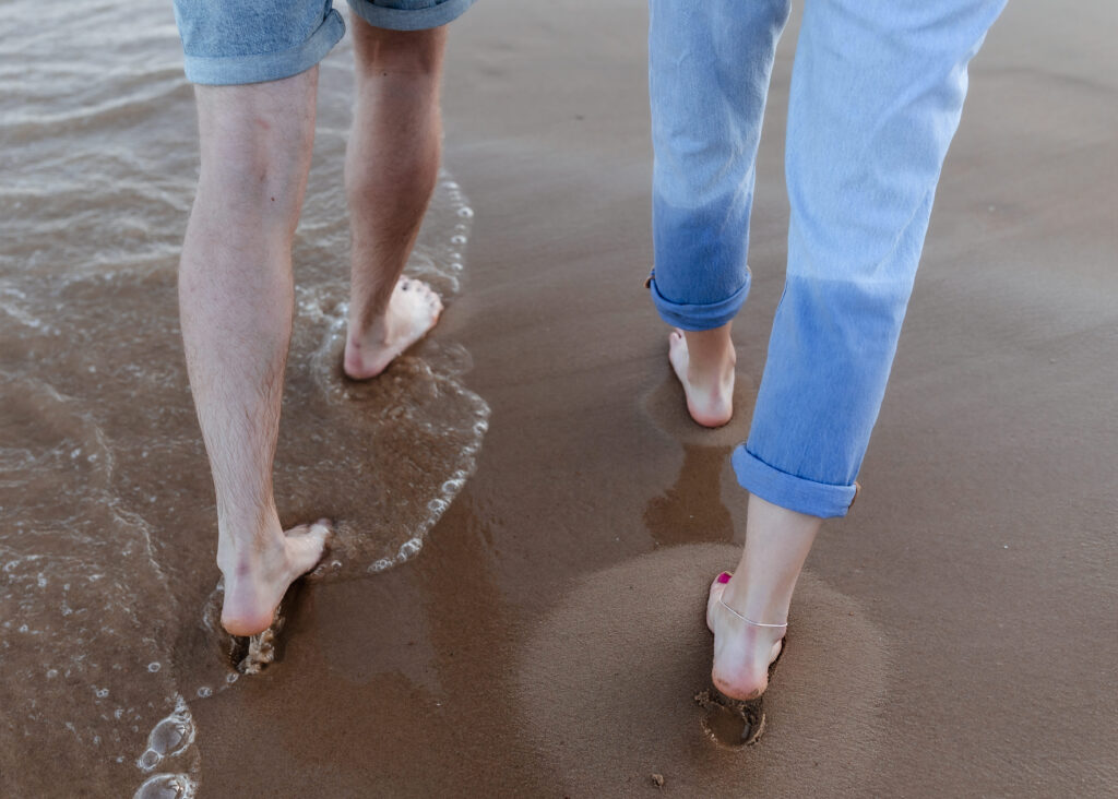 Two people walk barefoot on wet sand by the waters edge. One wears rolled-up jeans with painted toenails, the other wears shorts. Gentle waves touch their feet.