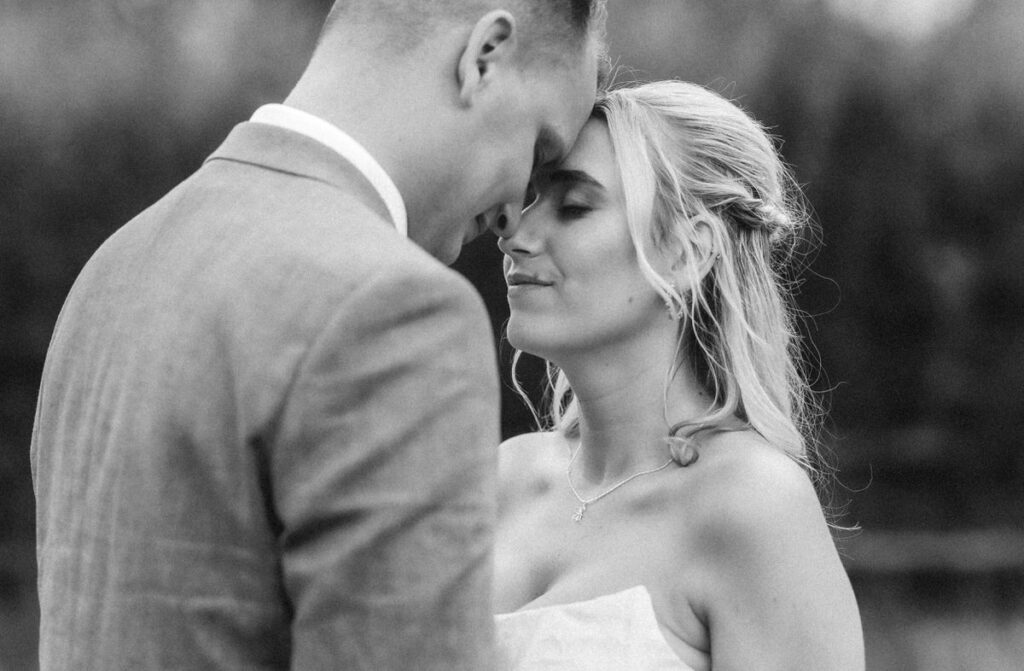 A couple stands close, heads touching gently. The woman, in a white dress, smiles softly with her eyes closed, while the man, in a suit, leans his forehead against hers. The moment is tender and intimate.