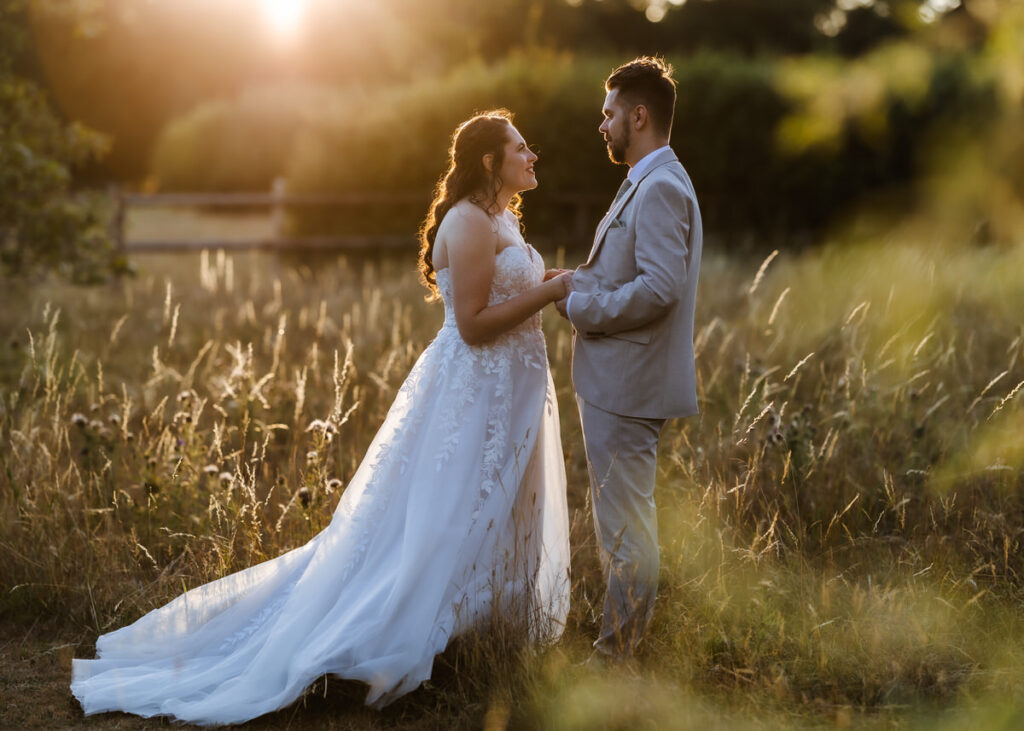 A bride in a white gown and a groom in a light suit stand holding hands in a sunlit grassy field at sunset, gazing into each others eyes.