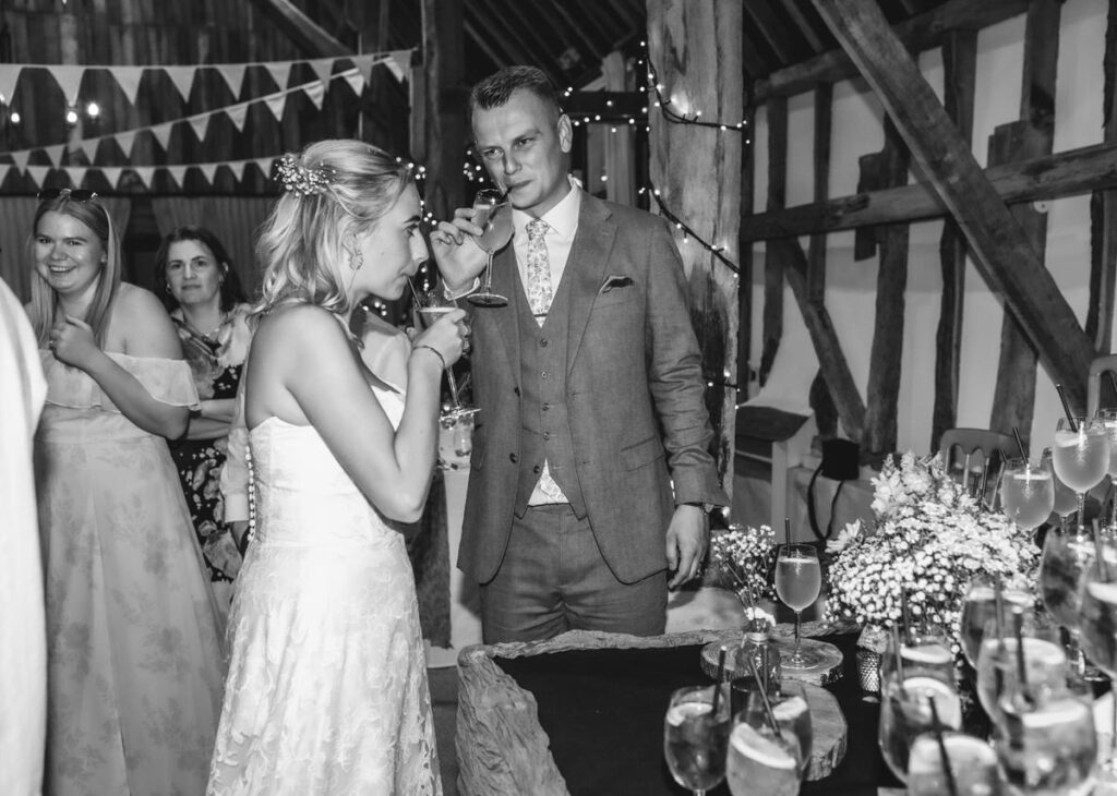 A bride and groom enjoy drinks at a rustic wedding reception with guests nearby, surrounded by string lights, bunting, and a table with flowers and glasses. The scene is lively and celebratory.