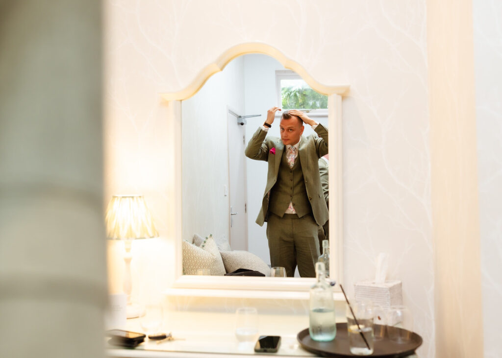 A man in a suit stands in front of a mirror, adjusting his hair. The scene includes a lit lamp, glass, water bottle, tissue box, and a tray on the table in front of the mirror.