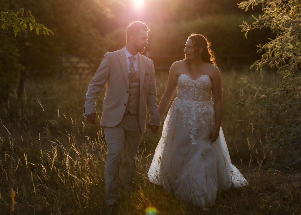 A bride and groom walk hand in hand through tall grass at sunset, both smiling and gazing at each other, bathed in warm, golden sunlight.