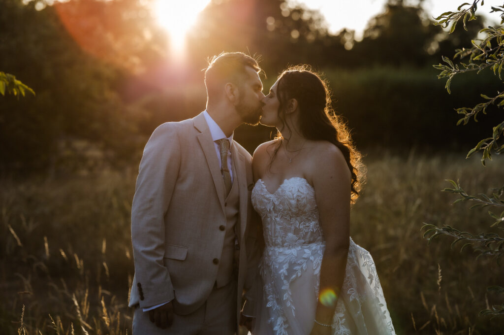 A bride and groom kiss outdoors at sunset, with golden light shining through trees and illuminating the brides lace wedding dress.
