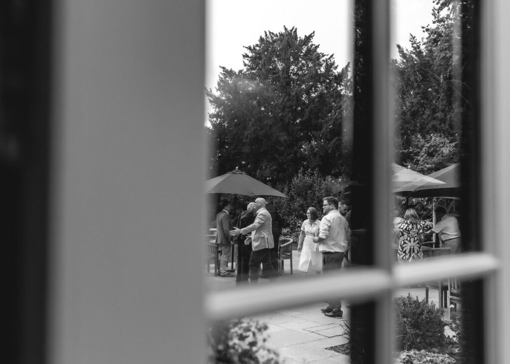 A black and white photo taken through a window shows people gathered outdoors, some under umbrellas, socializing in a garden setting. Trees and shrubs are visible in the background.