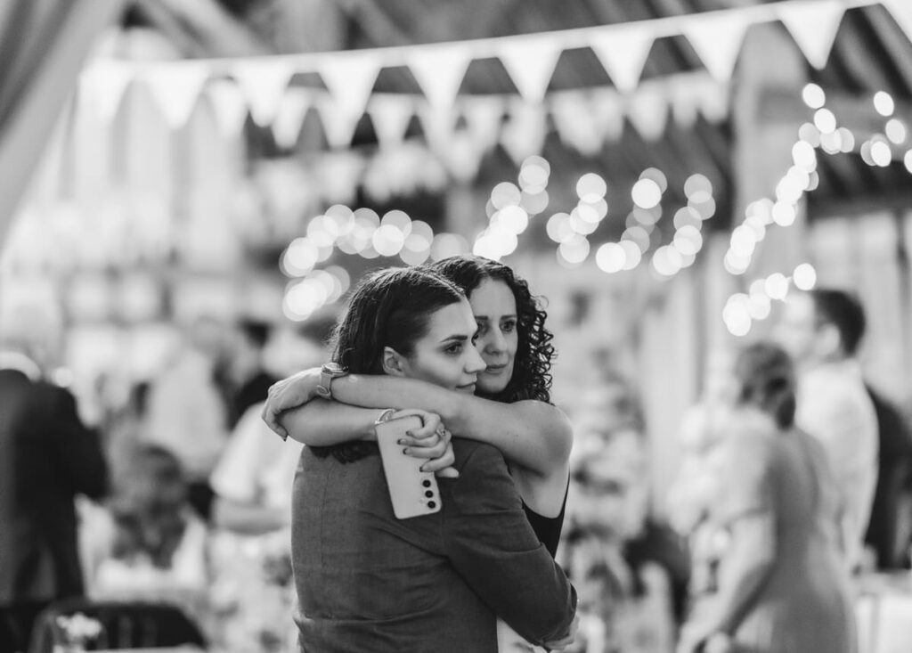 Two women embrace at a festive indoor event, with one holding a drink. String lights and bunting hang above a blurred crowd in the background. The image is black and white.