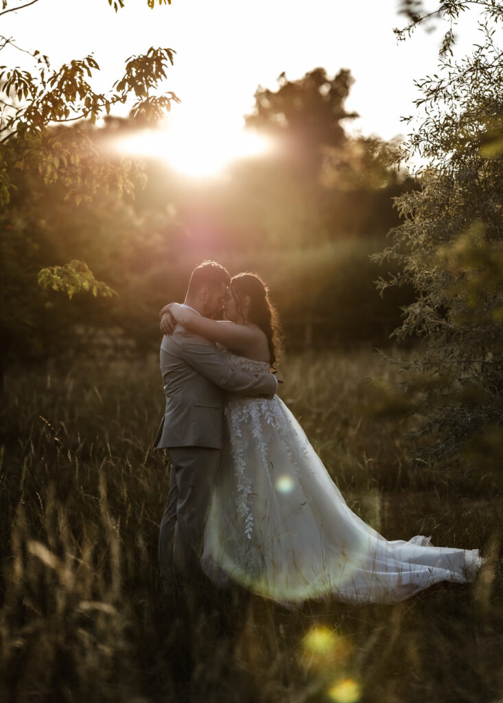 A bride and groom embrace in a grassy field at sunset, surrounded by trees, with sunlight creating a soft, romantic glow around them.