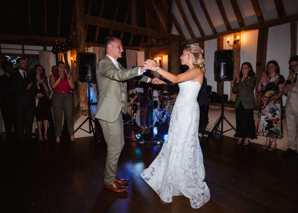 A bride and groom dance together in a rustic hall, smiling and holding hands, while guests watch and celebrate around them.
